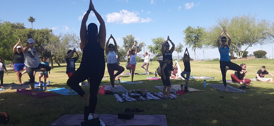 Photo shows a group of people in a yoga pose, standing on one leg with arms over their head, hands touching, in a local Laveen park.