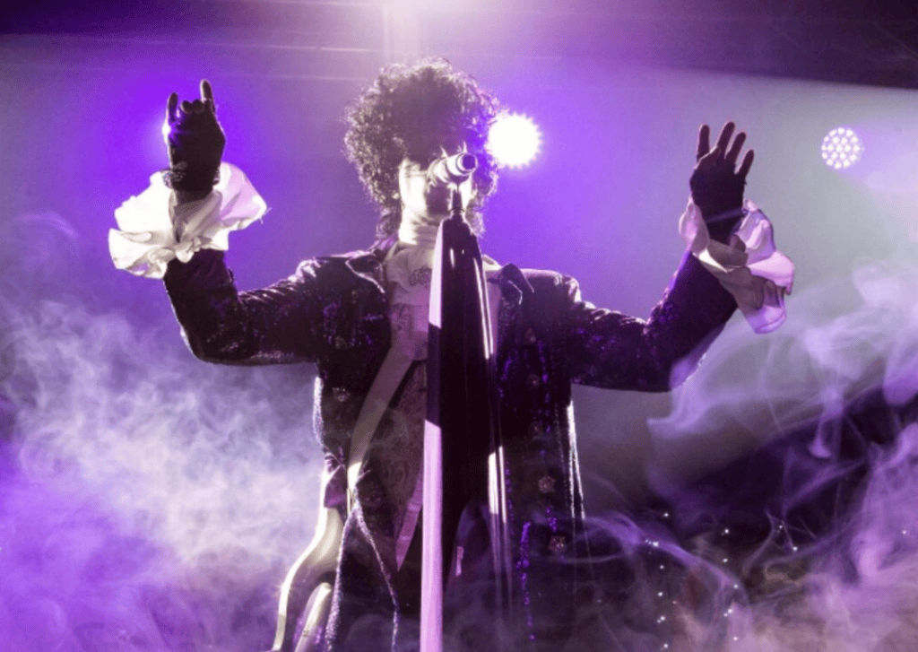 Prince tribute performer on stage beneath purple spotlights, dressed in a dark sequined jacket with white ruffled cuffs